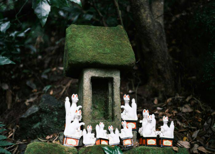 An unknown moss covered miniature shrine in the forest with small white fox statues surrounding the front.